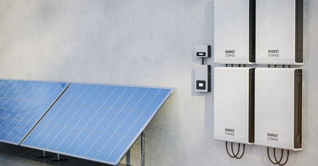 Several solar panels are angled against a concrete wall. Nearby, four energy storage panels are mounted on the wall.