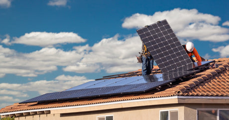 Two workers stand on the roof of a house. They carefully install several solar panels next to one another.