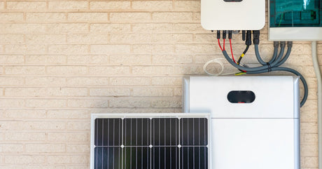 A large solar battery installed near a white brick wall. A small photovoltaic solar panel sits nearby.