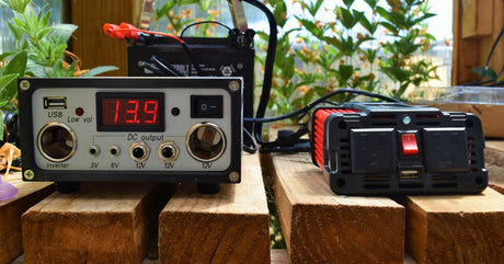A solar charge controller and power inverter are placed next to one another on a wooden bench. The controller reads 13.9.