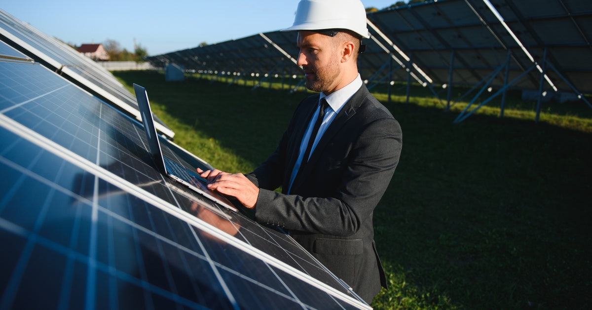 A man in a business suit and white hard hat stands outdoors by a large solar panel. He works on his laptop.