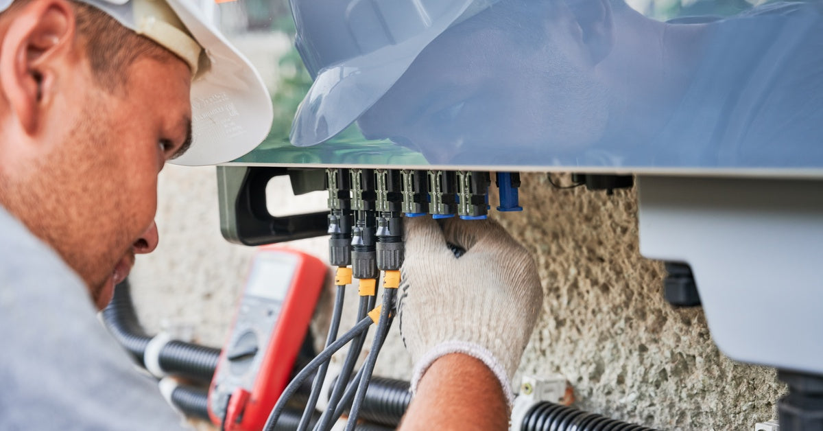 A technician wearing a white hard hat and gloves checks on the wiring inside an electrical inverter box.
