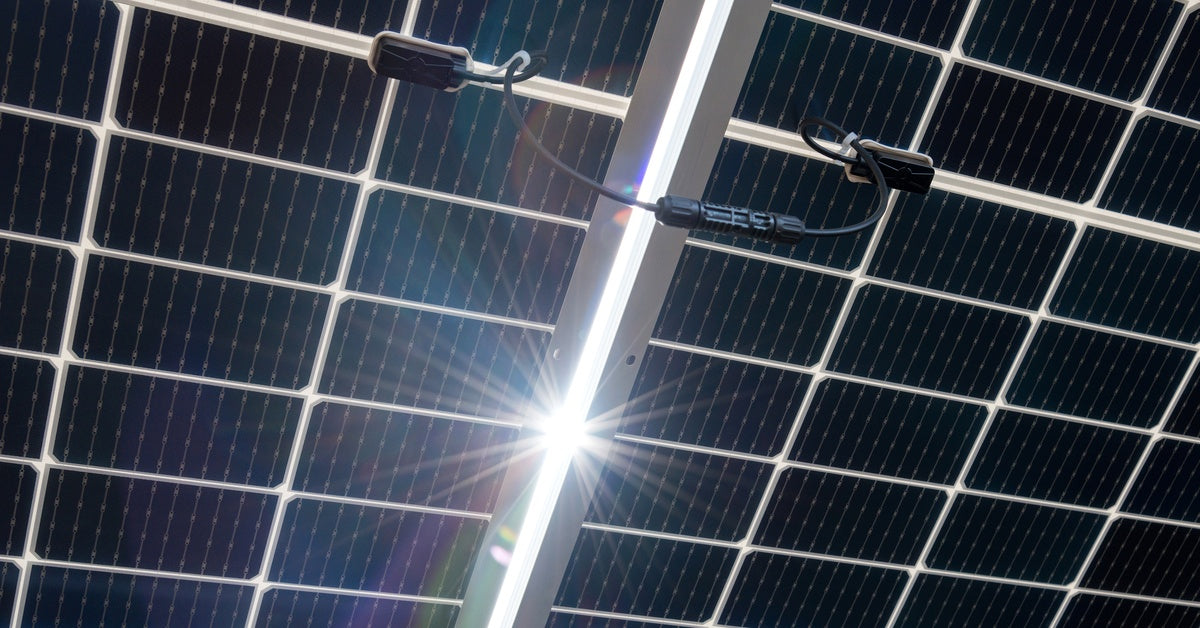 The underside of a large set of bifacial solar panels is visible. The sun peeks through from between two panels.