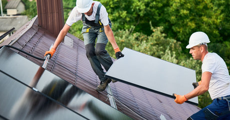 Two workers in white hard hats climb a roof to install solar panels. Both of them hold onto a single panel as they climb.