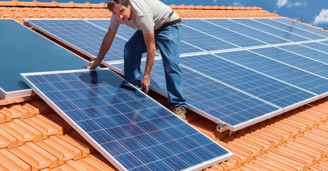 A man in a t-shirt and jeans stands on the angled roof of a house. He pulls one large solar panel into position.